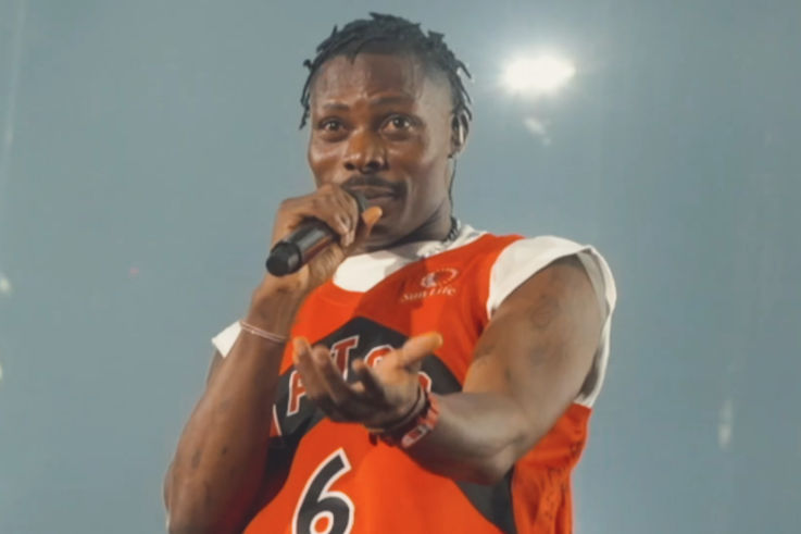 Asake in an orange Raptors jersey singing into a microphone on stage in Toronto