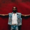 Black Sherif stands with arms outstretched against a vivid red backdrop, wearing a denim vest over a white shirt.