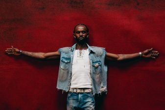 Black Sherif stands with arms outstretched against a vivid red backdrop, wearing a denim vest over a white shirt.