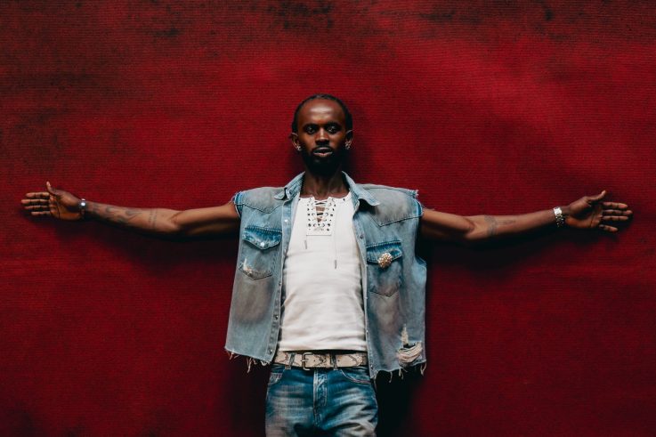 Black Sherif stands with arms outstretched against a vivid red backdrop, wearing a denim vest over a white shirt.