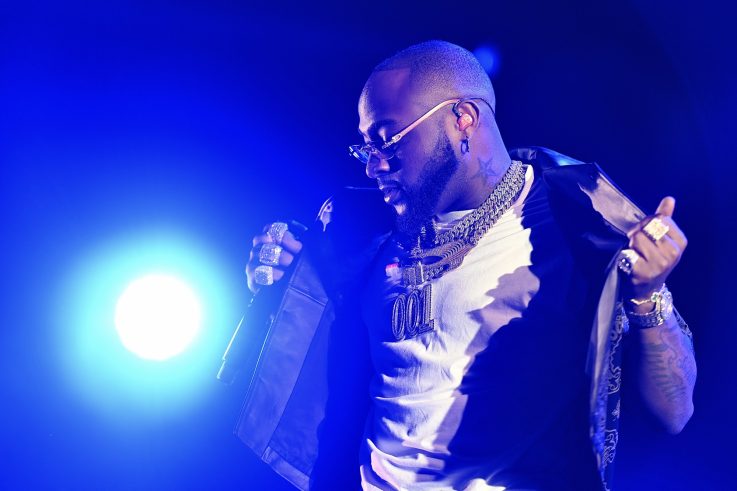Davido performing on stage in a 001 chain and white T-shirt under blue lights at Reggae Geel in 2022