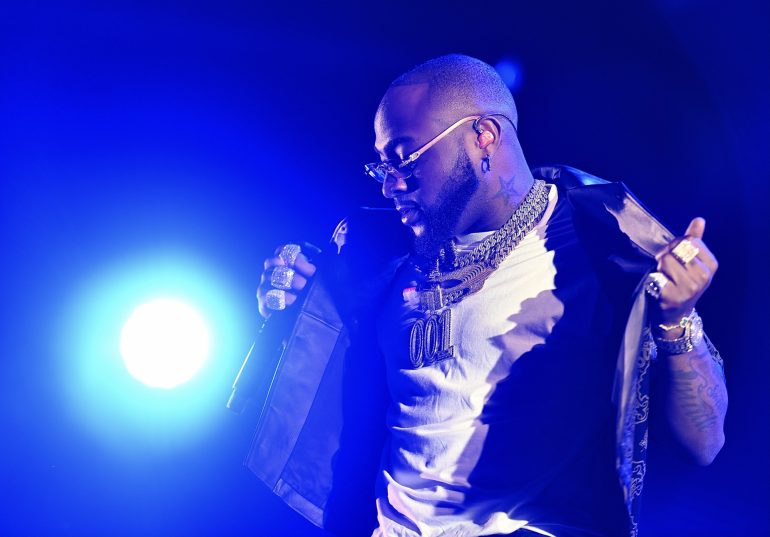 Davido performing on stage in a 001 chain and white T-shirt under blue lights at Reggae Geel in 2022