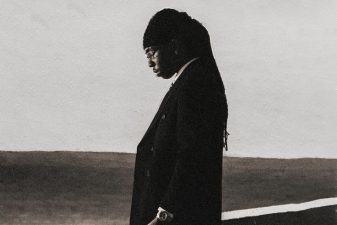 FOLA "9" album cover: FOLA in a dark suit with his head bowed against a pale sky, a gothic "9" in the corner.
