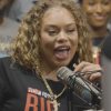 Latto with curly blonde hair and a gold chain smiling into a studio microphone in a Hot 107.9 T-shirt