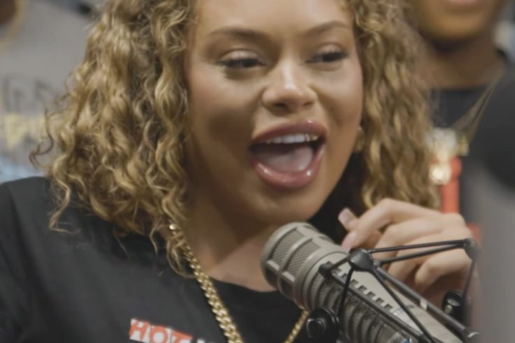 Latto with curly blonde hair and a gold chain smiling into a studio microphone in a Hot 107.9 T-shirt