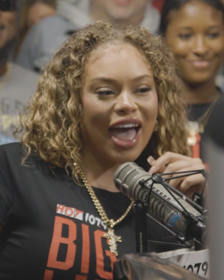 Latto with curly blonde hair and a gold chain smiling into a studio microphone in a Hot 107.9 T-shirt