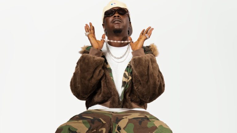 Magicsticks in a camouflage jacket and cream cap, hands raised